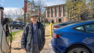 Harvey Meer, wearing a fall jacket and hat, proudly stands in front of an electric vehicle charging station on a crisp fall day. Two electric vehicles are charging behind him.